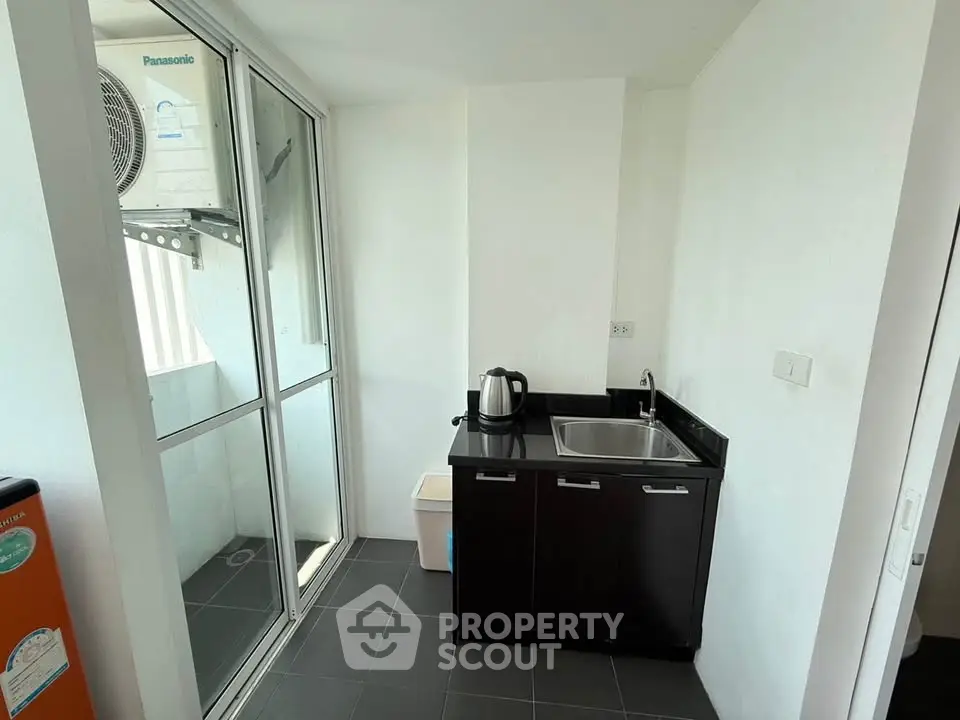 Compact utility area with sink and kettle, featuring a glass door and air conditioning unit.