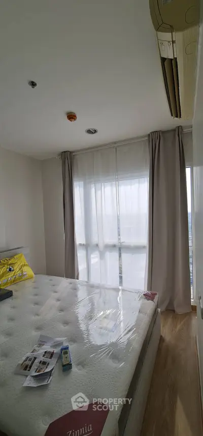 Bright bedroom with large window and air conditioning unit, featuring a comfortable bed and wooden flooring.