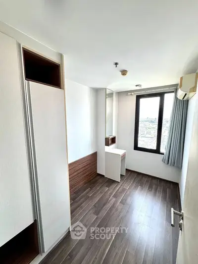 Modern minimalist bedroom with wooden flooring and large window offering city views.