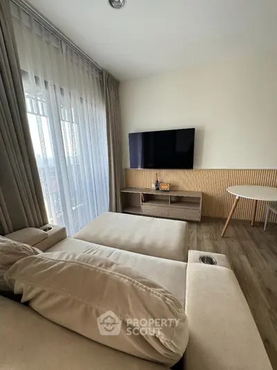 Modern living room with cozy beige sofa and wall-mounted TV, featuring floor-to-ceiling windows.