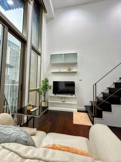 Modern living room with high ceiling and staircase, featuring a cozy sofa and large windows for natural light.