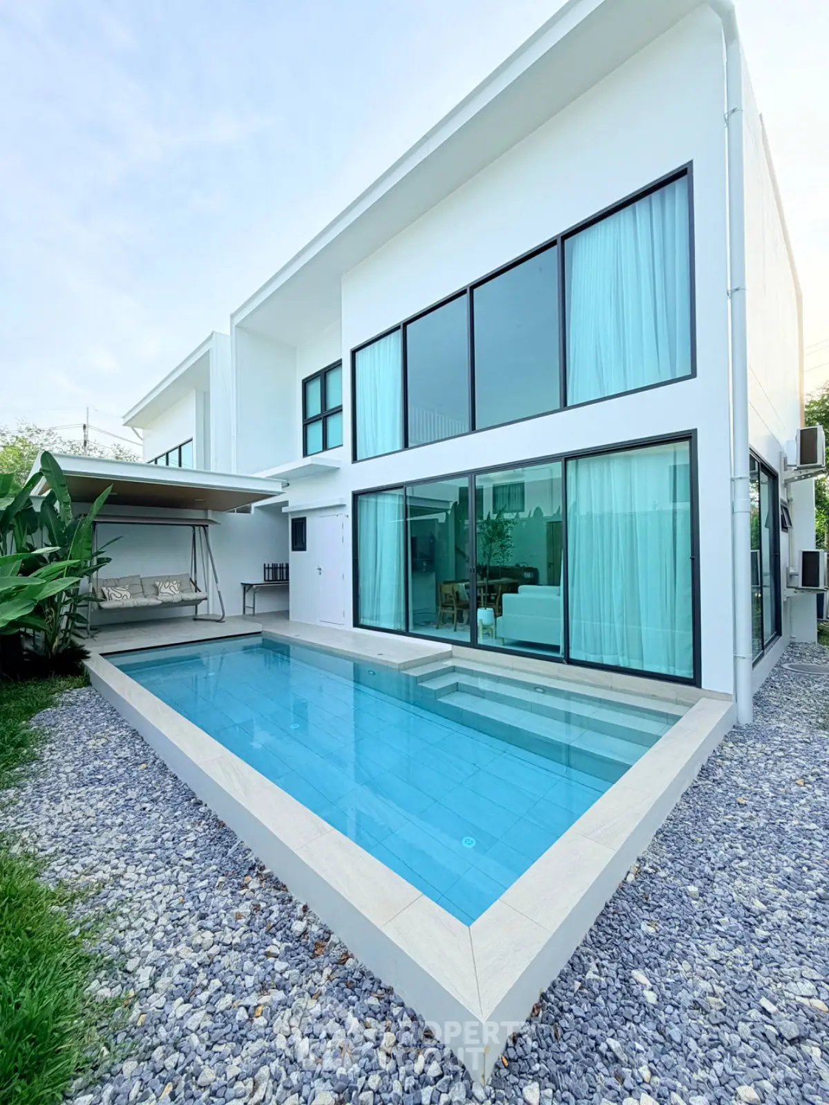 Modern white house with large windows and a sleek outdoor pool, surrounded by a gravel landscape.