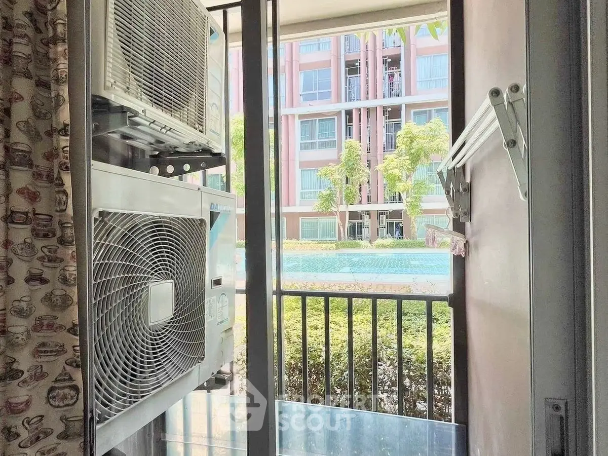 Charming balcony view with air conditioning units overlooking lush greenery and modern building facade.