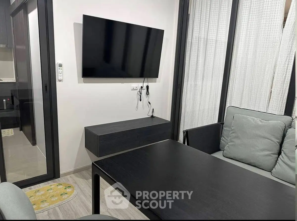 Modern living room with sleek black furniture and wall-mounted TV, featuring large windows with sheer curtains.