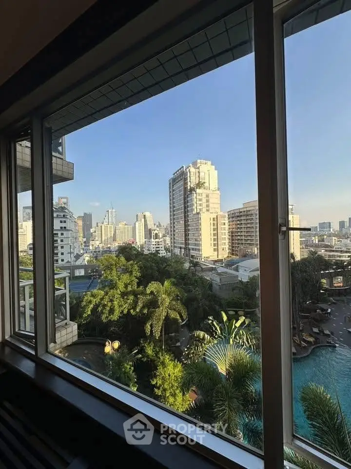 Stunning cityscape view from a high-rise window overlooking lush greenery and a sparkling pool.