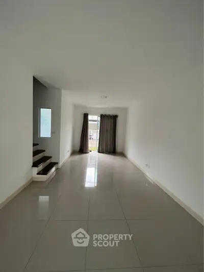 Spacious and bright living room with glossy tiled flooring, large windows draped with elegant curtains, and a modern staircase leading to an upper level.