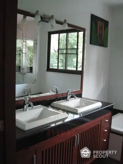 Elegant dual-sink bathroom vanity with rich wooden cabinetry and modern fixtures, set against a serene backdrop of lush greenery visible through the window.