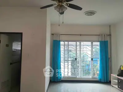 Spacious living room with ceiling fan and large window, featuring blue curtains and ample natural light.