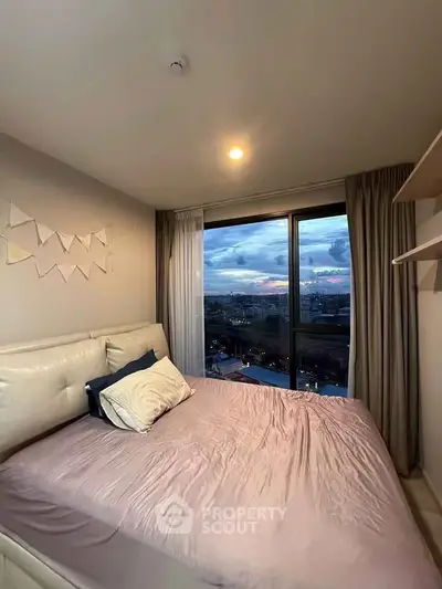Cozy bedroom with large window offering stunning city view at sunset.