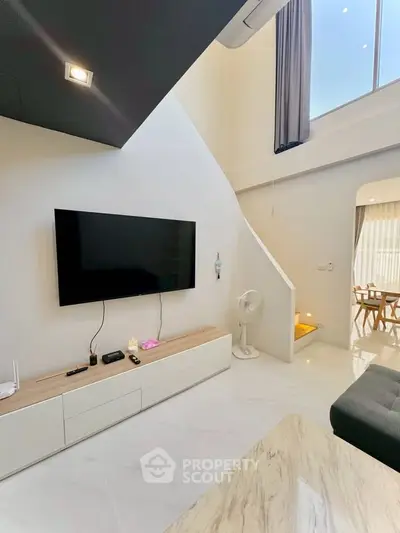 Modern living room with high ceiling and large TV, featuring sleek white furniture and natural light.