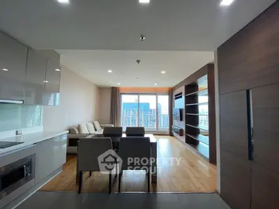 Spacious modern living room with open kitchen and city view, featuring sleek furniture and wooden flooring.