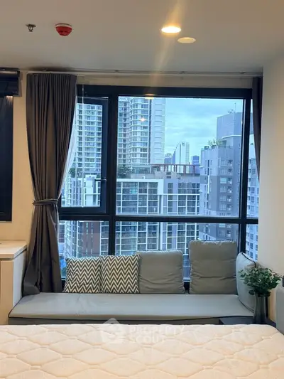 Modern bedroom with large window offering stunning city view, cozy seating area, and elegant decor.