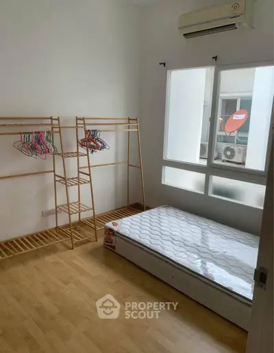 Minimalist bedroom with wooden floor, mattress, and clothes rack in bright natural light.