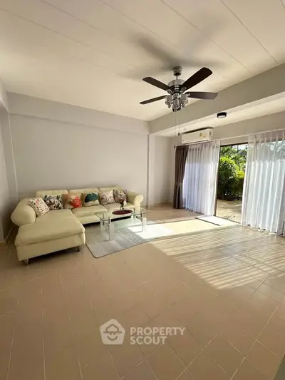 Spacious living room with natural light and garden view, featuring a cozy sectional sofa and ceiling fan.