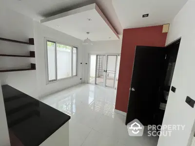 Spacious modern kitchen with glossy countertops, ample natural light, and a striking red accent wall leading to a bright living area.