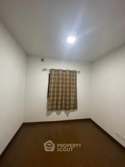 Minimalist empty room with wooden flooring and a single window with curtains.