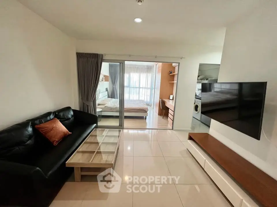 Modern living room with sleek black sofa, glass coffee table, and wall-mounted TV, leading to a cozy bedroom.