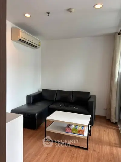 Modern living room with black leather sofa and air conditioning, featuring wooden flooring and natural light.