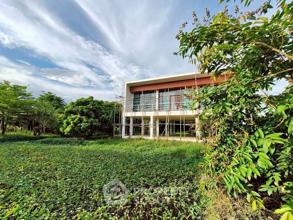 Modern two-story house with large windows surrounded by lush greenery and open sky.