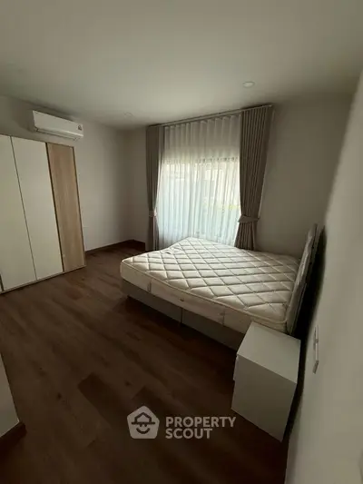 Spacious bedroom with wooden flooring and natural light, featuring a large window and modern air conditioning.