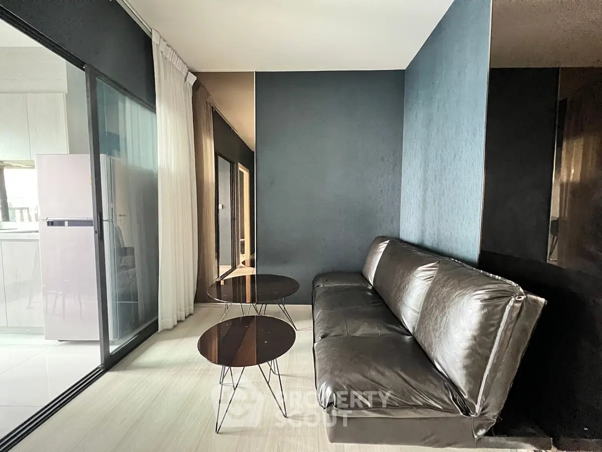 Modern living room with sleek black leather sofa and glass coffee tables, adjacent to a bright kitchen area.