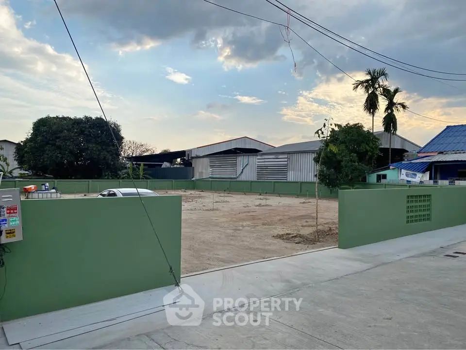 Spacious vacant lot with green fencing, ideal for development, surrounded by industrial buildings and clear skies.