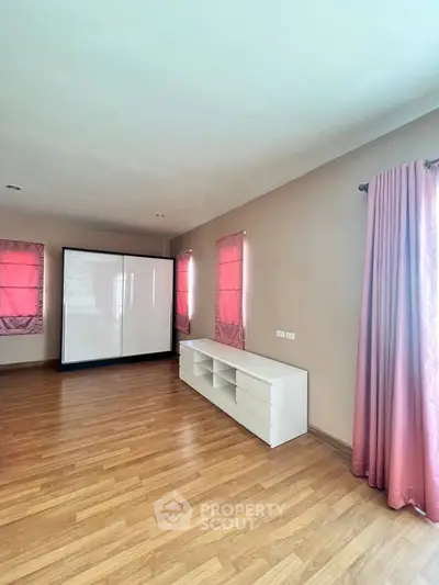 Spacious bedroom with wooden flooring and pink curtains, featuring a sleek white wardrobe.