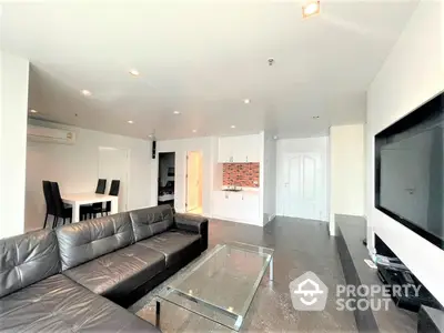 Spacious living room with sleek black leather sofa, glass coffee table, and modern entertainment unit, adjacent to an open-plan kitchen with chic brick backsplash.