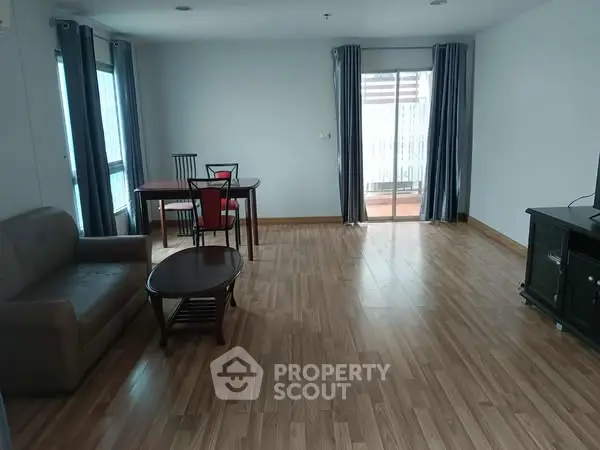Spacious living room with wooden flooring and modern furniture, featuring large windows and balcony access.