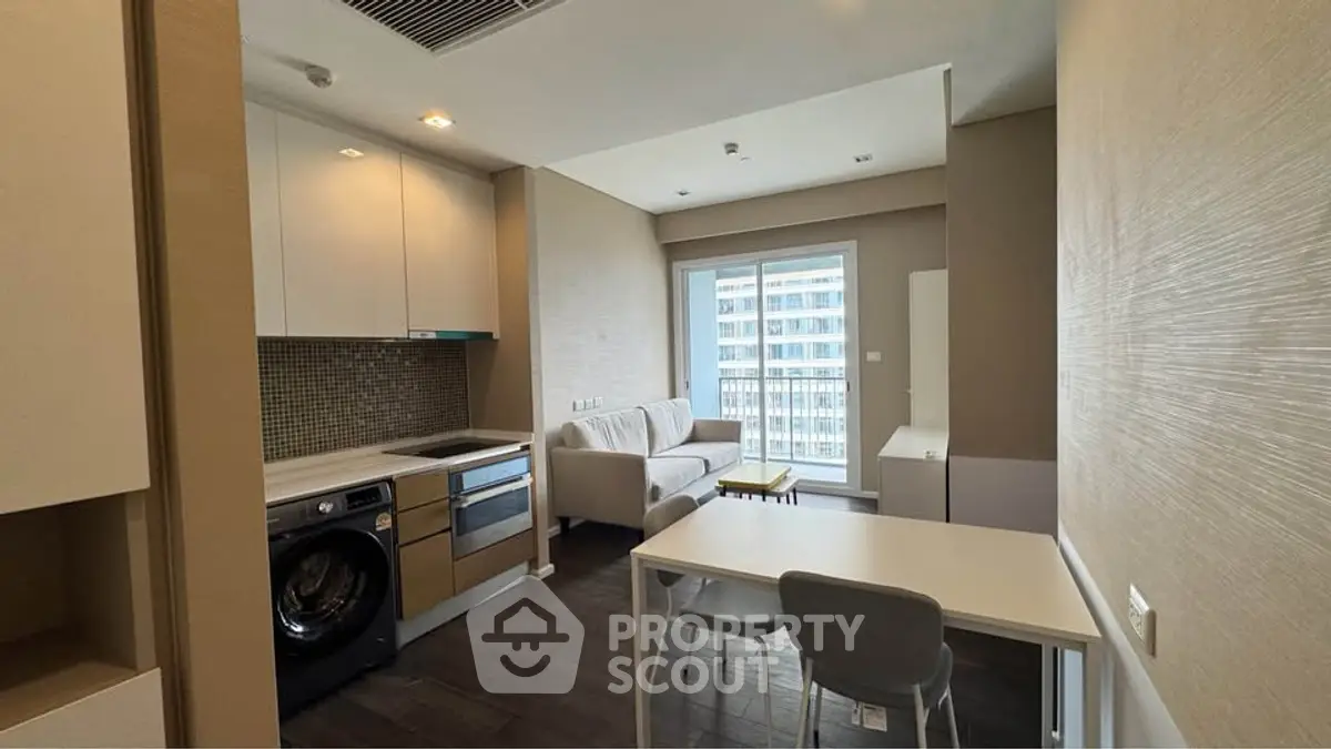 Modern apartment interior with open kitchen and cozy living area, featuring a washing machine and large window view.