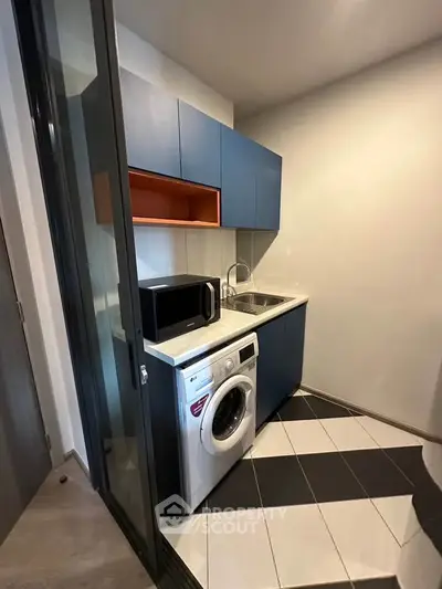 Compact modern kitchen with blue cabinets, washing machine, and microwave in a sleek apartment.