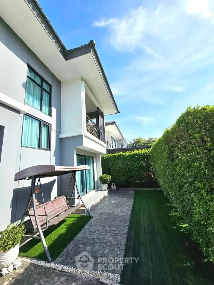 Modern two-story house with lush garden and cozy swing under clear blue sky.