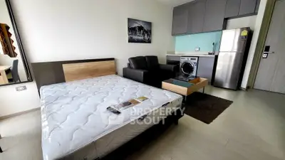Modern studio apartment with bed, sofa, and compact kitchen featuring washing machine and fridge.
