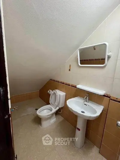 Compact bathroom with sloped ceiling, featuring a toilet, sink, and mirror.