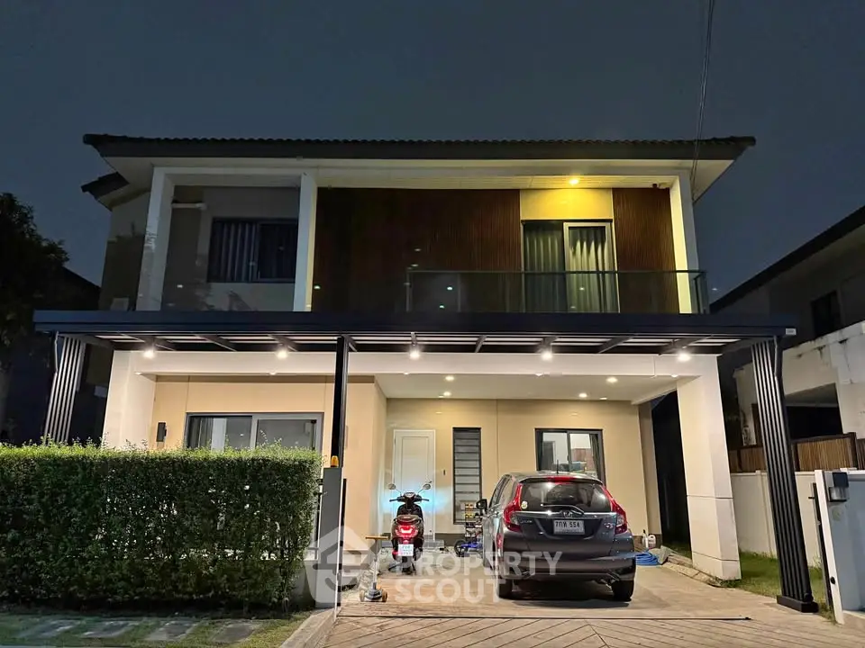 Modern two-story house with carport and balcony, illuminated at night, showcasing contemporary design and spacious driveway.