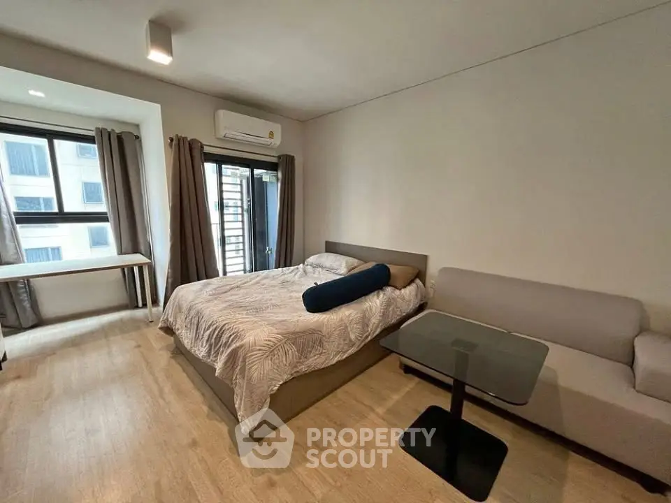 Modern studio apartment with cozy bed, sofa, and natural light from large window.