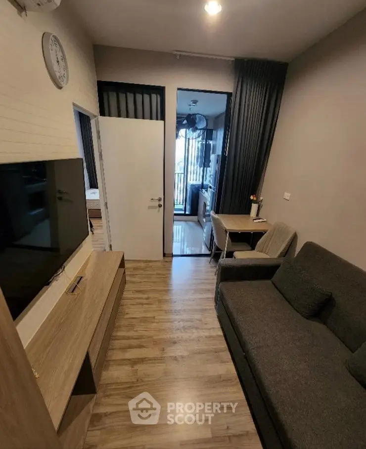 Modern living room with wooden flooring, cozy sofa, and wall-mounted TV leading to a bright balcony.