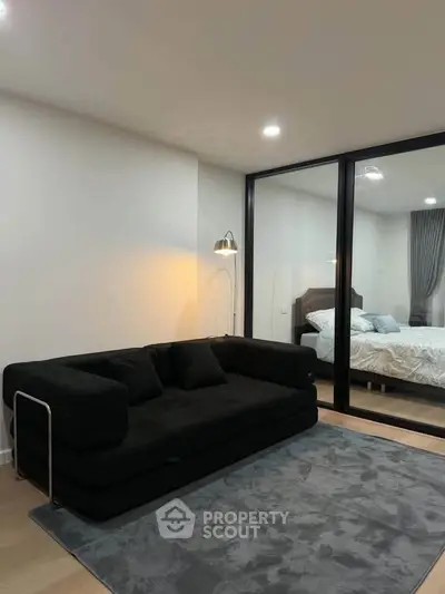 Modern living room with sleek black sofa and glass partition leading to a cozy bedroom.