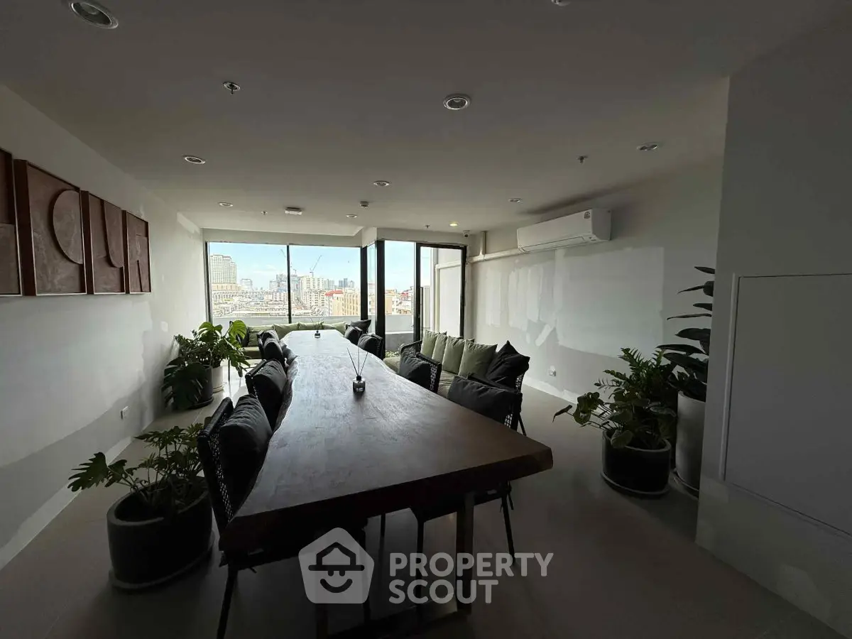 Spacious modern living room with large windows and city view, featuring elegant dining table and lush plants.