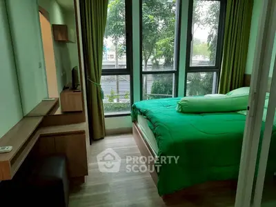 Cozy bedroom with large windows and green bedding, offering a serene view of lush trees.
