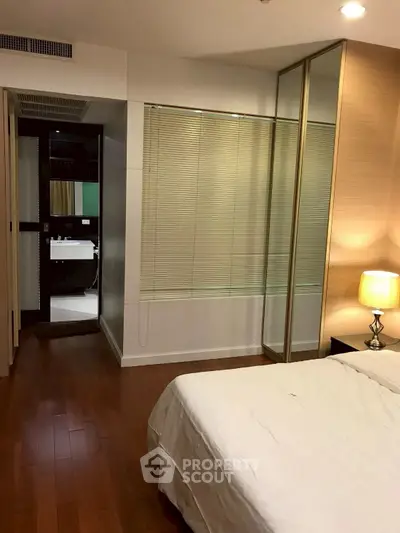 Modern bedroom with wooden flooring and mirrored closet doors, adjacent to a sleek bathroom.