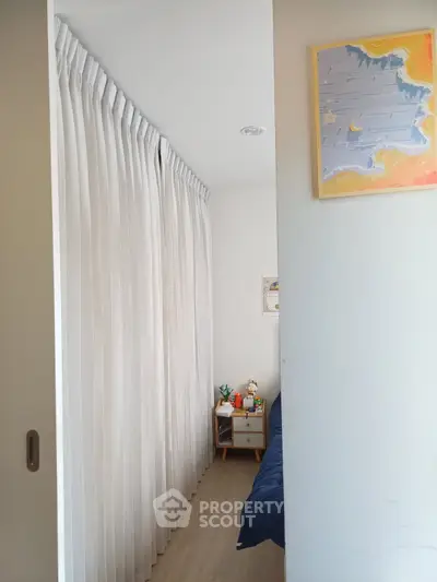 Cozy bedroom with white curtains and wooden flooring, featuring a small bedside table and colorful wall art.
