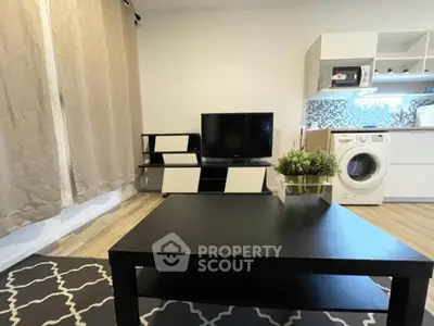 Modern living room with TV, stylish furniture, and compact kitchen area featuring washing machine.