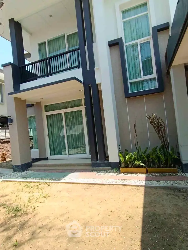 Modern two-story house with large windows and balcony, featuring a small garden area.