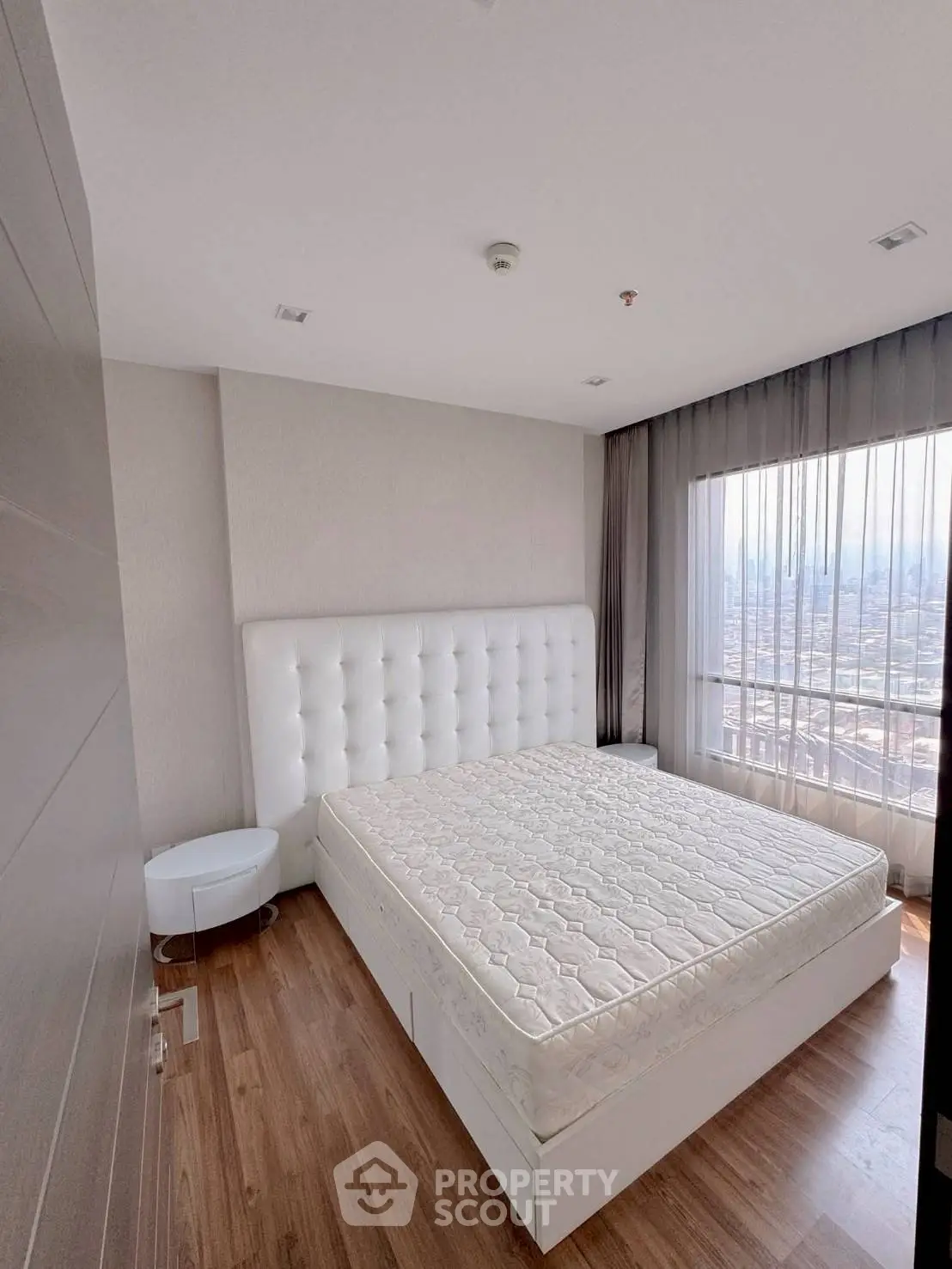 Spacious bedroom with large window offering stunning city views, featuring a modern white bed and wooden flooring.