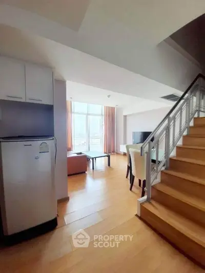 Spacious modern living room with wooden flooring and staircase, featuring large windows for natural light.