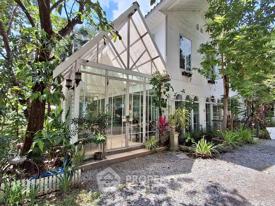 Charming white house with glass sunroom surrounded by lush greenery and gravel pathway.