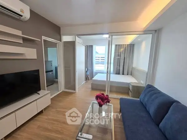 Modern open-plan living room with glass partition to bedroom, featuring sleek furniture and wooden flooring.
