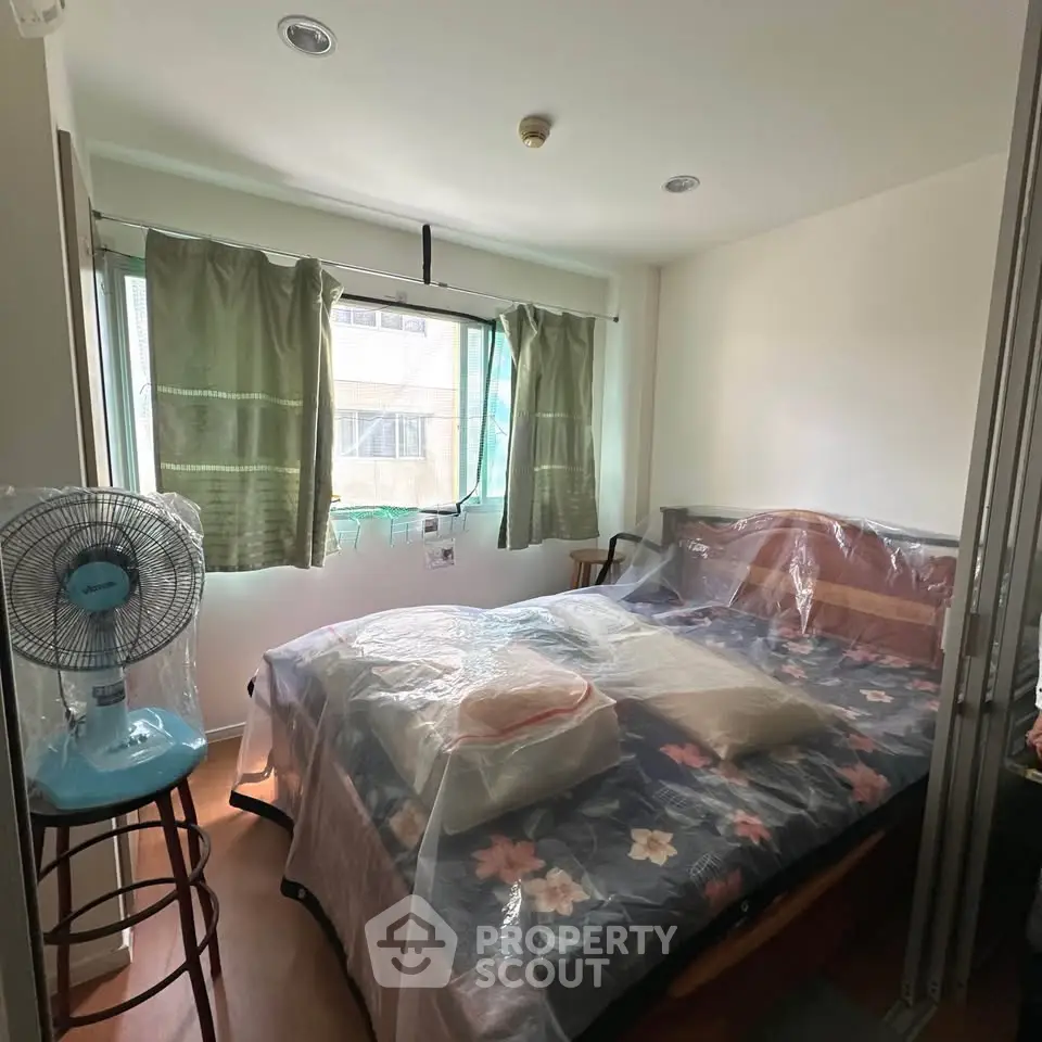 Cozy bedroom with large window and floral bedding, featuring a standing fan and natural light.