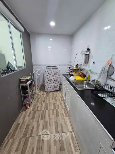 Compact kitchen with washing machine and dish racks, featuring tiled flooring and modern lighting.